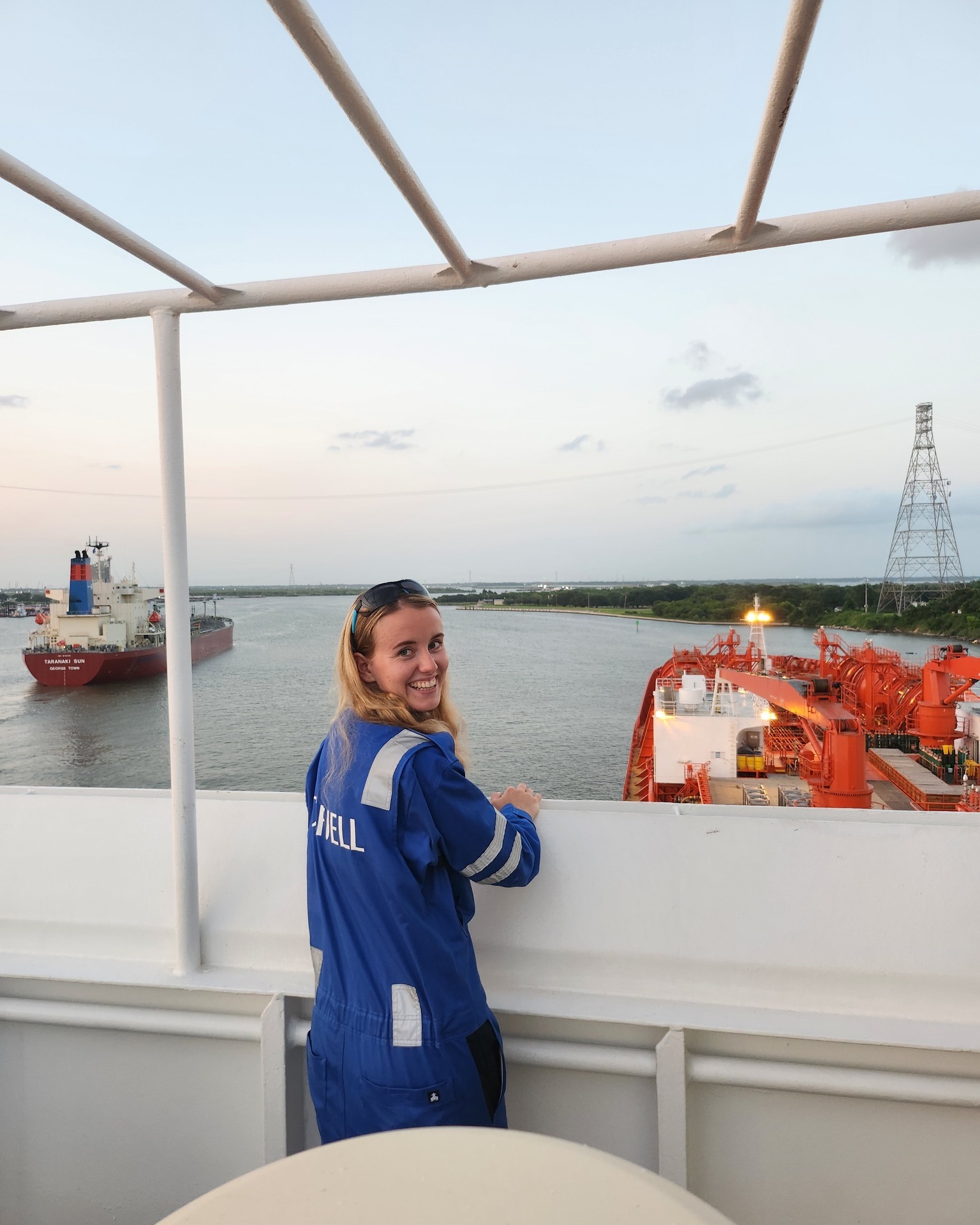 A female cadet stands on deck in Odfjell workwear, smiling as she looks out over the waterway and nearby vessels.