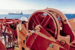 Odfjell crew member operating winch system on chemical tanker with cargo ship in background.