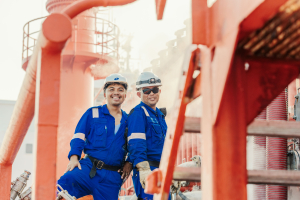 Two Odfjell seafarers in blue uniforms and safety helmets smiling on deck surrounded by red ship structures and steam.