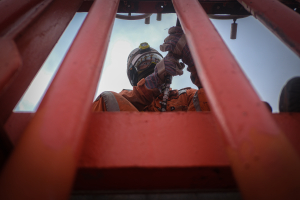 Crew member in orange safety gear performing maintenance work through red ship railing.
